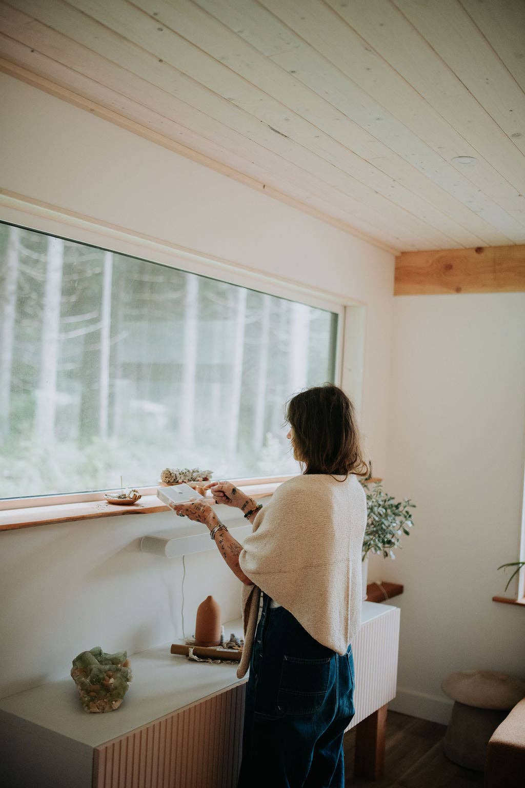 Woman wearing a beige shawl in a cozy room with large windows, looking out at a forest.