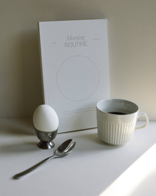 White mug with coffee, egg, spoon, and 'The Morning Routine Journal' book on a light surface.